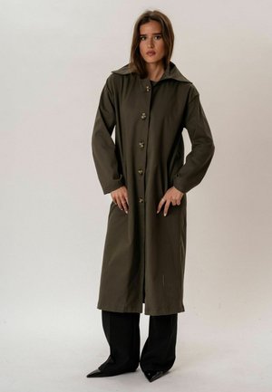 Femme aux cheveux bruns portant un long manteau boutonné vert olive par-dessus un pantalon noir et des chaussures noires pointues, debout devant un fond uni.