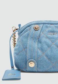 Hellblaues Denim-Päuschen mit gestepptem Muster, Hardware in Goldtönen und einem abnehmbaren Anhänger. Verfügt über einen geflochtenen Kettenriemen und Logodetails.