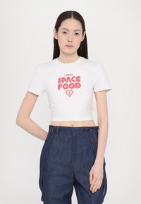 Bijela skraćena majica s crvenim grafičkim tekstom "Fiorucci SPACE FOOD" i malim srcem. Uparena s tamnoplavim trapericama.