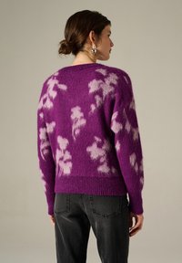 Pull violet et duveteux avec des motifs floraux roses. Présente un col rond et un ourlet côtelé. Porté avec un jean foncé.