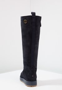 Bota de ante negra hasta la rodilla con una textura suave, panel lateral elástico, detalle de hardware dorado redondo y suela de goma con un ligero relieve.