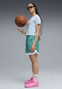 Camiseta corta de color azul claro con un gráfico, pantalones cortos con estampado en tonos verdes, zapatillas deportivas rosas y gafas de sol, sosteniendo un balón de baloncesto.