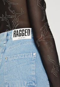 Traperice kratke hlače svijetlo plave boje s bijelom oznakom pod nazivom "RAGGED JEANS", uparenim s crnom prozirnom majicom s metalnim ukrasima.