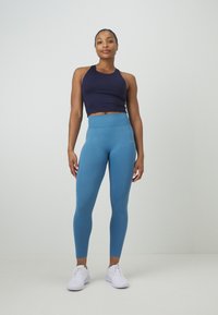 Top corto navy con scollatura a halter abbinato a leggings teal a vita alta e texture. Indossato con scarpe da ginnastica bianche su uno sfondo neutro.