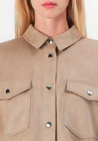 Beige suède shirt met een kraag, zilveren drukknopen en twee borstzakken. De textuur is zacht en mat.