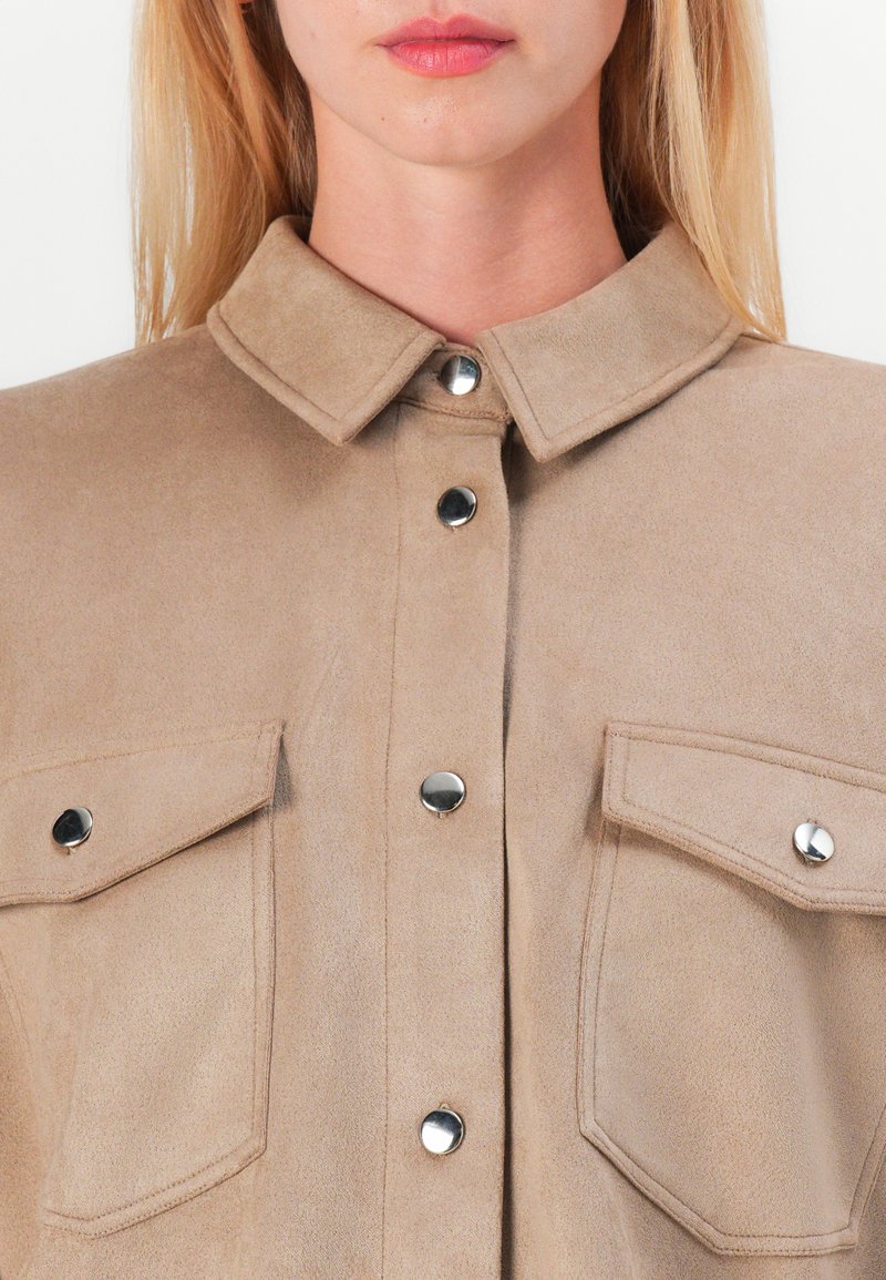 Camicia in suede beige con colletto, bottoni a pressione argentati e due tasche sul petto. La texture è morbida e opaca.