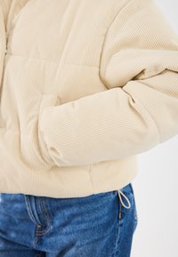 Kremo spalvos korduroy švarko rankovė su ranka kišenėje, derinama su mėlynomis džinsinėmis kelnėmis prieš šviesų vientisą foną.