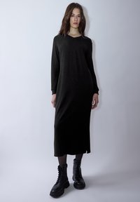 Lange schwarze Kleid mit V-Ausschnitt, langen Ärmeln und seitlichen Schlitzen. Dazu schwarze Schnürstiefel im Combat-Stil. Glatte Textur, minimalistisches Design.