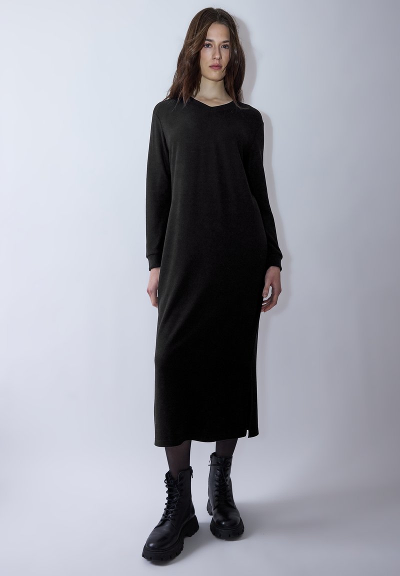 Lange schwarze Kleid mit V-Ausschnitt, langen Ärmeln und seitlichen Schlitzen. Dazu schwarze Schnürstiefel im Combat-Stil. Glatte Textur, minimalistisches Design.