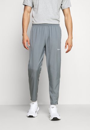 Mand iført grå Nike træningsbukser og hvide Nike sneakers står foran en ensartet lys baggrund.