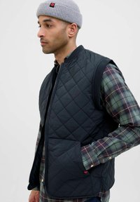 Gilet trapuntato blu navy con chiusura a zip, dotato di una grande tasca frontale e aperture per le braccia in costina, indossato sopra una camicia a quadri con tonalità verdi e blu.