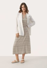 Blazer blanco de doble botonadura sobre un vestido beige con estampado. El vestido es de longitud midi y tiene escote en V. Sandalias deslizantes beige completan el atuendo.