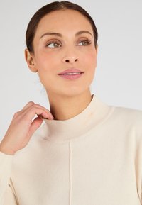 Maglione a collo alto in crema realizzato in morbido tessuto lavorato a maglia. Presenta un colletto a coste e un sottile design testurizzato, aderendo delicatamente al corpo.