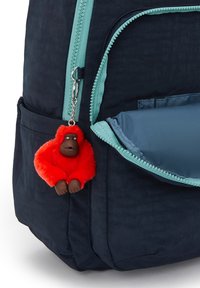 Námořnický batoh s tyrkysovým zipem a kapsou, který obsahuje červený chlupatý klíčenku ve tvaru gorily s hnědým obličejem.