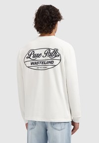 Witte longsleeve met zwarte tekst op de achterkant: "Pure Path WASTELAND." Ronde hals, losse pasvorm, zachte katoenen structuur.