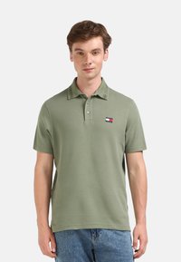 Polo verde oliva con cuello y tres botones, que presenta un pequeño parche de logo en el lado izquierdo del pecho. Combinado con jeans azules claros.