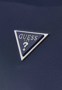 Jūros mėlynos spalvos audinys su trikampiu sidabriniu metalo logotipu, kuriame yra "GUESS USA" ir klaustukas. Glotnus tekstūra ir elegantiškas dizainas.