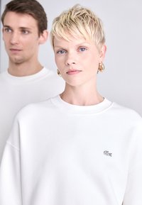Weißer Sweatshirt mit Rundhalsausschnitt und gesticktem Logo. Der Stoff hat eine glatte Textur. Zwei Modelle sind im Hintergrund.