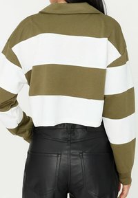 Camisa corta a rayas en verde oliva y blanco con cuello, mostrada por la parte de atrás, combinada con pantalones negros de cintura alta. Textura de tela suave.