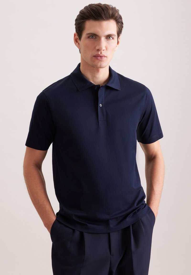 Jeune homme à la peau claire et aux cheveux bruns portant un polo bleu marine foncé et un pantalon assorti, les mains posées décontractées dans les poches, fond neutre.