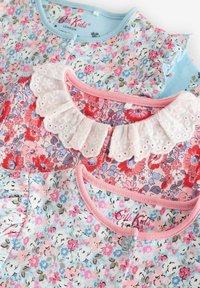 Zwei Kinderblusen mit Blumenmuster, eine in Hellblau mit kleinen pinken Blumen und Rüschen, die andere in Pink mit einem Spitzenkragen und lebhaftem Blumenmuster.