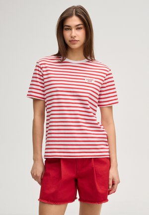 Kobieta ubrana w czerwono-białą poziomo paskowaną koszulkę z logo "JOOP! JEANS" oraz czerwone, postrzępione szorty, stojąca na jednolitym tle.