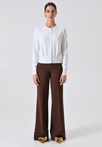 Una donna in piedi indossa un cardigan bianco con bottoni, pantaloni larghi marroni con risvolti e scarpe dorate a punta con grandi fiocchi.