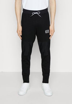 Homme portant un pantalon de jogging noir avec des patches aux genoux à l'avant, un cordon blanc, le logo EA7 sur la cuisse, des baskets blanches et un t-shirt blanc.