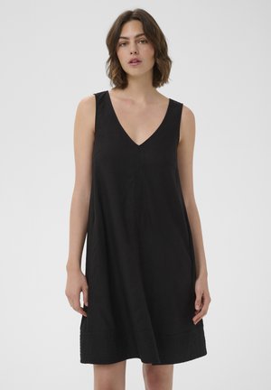 Mujer con cabello castaño hasta los hombros, vestida con un vestido negro sin mangas con escote en V, de pie frente a un fondo claro y liso.