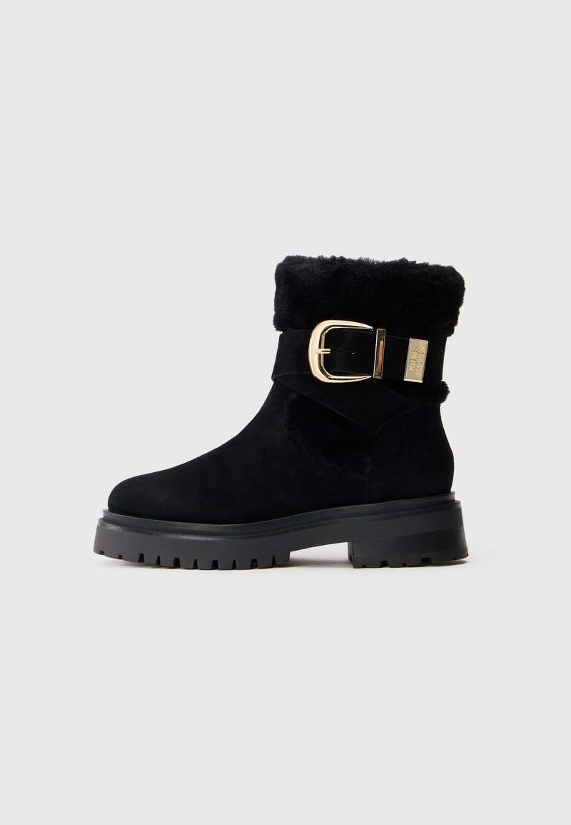 Botine negre din suede, cu talpă groasă din cauciuc, cu mansetă din blăniță pliată și detaliu de curea lată cu cataramă aurie.
