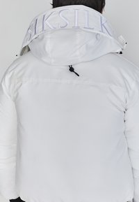 Hvid pufferjakke med hætte, der har en høj krave og broderet logo på ryggen. Materialet er glat og polstret for isolering.
