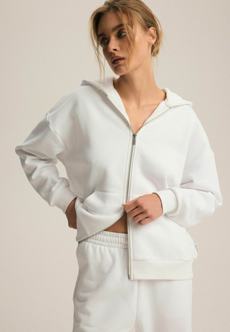 Biały rozpinany hoodie wykonany z mieszanki bawełny, charakteryzujący się luźnym krojem, kieszeniami kangurkami oraz ściągaczami przy mankietach. Połączony z dopasowanymi spodniami ze sznurkiem.