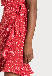 Vestido rojo con lunares, con dobladillo con volantes, detalle de lazo en el costado y tela texturizada. La parte visible incluye el brazo y la mano.