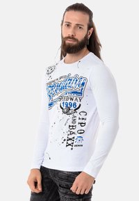 Weißes Langarm-T-Shirt mit grafischen Drucken in Blau und Schwarz, ausgestattet mit Spritzdetails und Schriftzug, getragen mit dunkler Jeans.