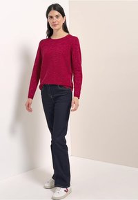 Röd marmoriserad tröja, rund halsringning, kombinerad med mörka jeans. Vita sneakers med röda detaljer. Minimalistisk bakgrund.