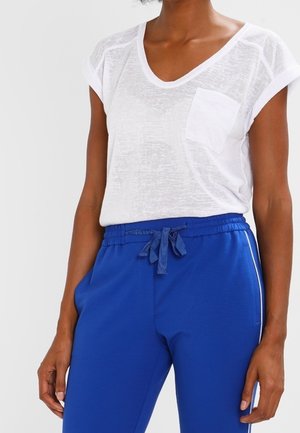 Camiseta blanca de manga corta con cuello en V y bolsillo, combinada con pantalones azules con cordón, rayas laterales y ajuste holgado.