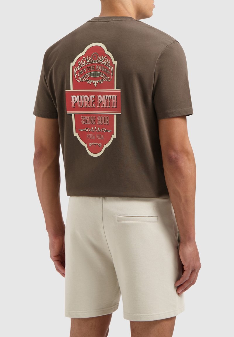 Bruine katoenen T-shirt met een opvallende rode en crèmekleurige grafische afbeelding op de achterkant met "PURE PATH" en decoratieve elementen. Draagt met beige shorts.
