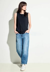 Top nero senza maniche con finitura testurizzata, abbinato a jeans chiari a vita alta e sneakers bianche. Lo sfondo semplice crea contrasto.