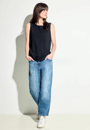 Top sans manches noir avec une finition texturée, associé à un jean large bleu clair et des baskets blanches. Un fond uni ajoute du contraste.