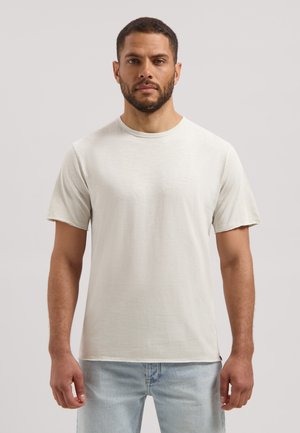 Man met kort haar en baard, gekleed in een eenvoudig lichtbeige T-shirt met korte mouwen en lichtblauwe jeans, staand tegen een effen lichte achtergrond.