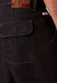 Κοντινή λήψη στην πίσω τσέπη μαύρου τζιν από denim με καπάκι, αντίθετη πορτοκαλί ραφή και καφέ δερμάτινο μπάλωμα με το λογότυπο Calvin Klein πάνω από την τσέπη.