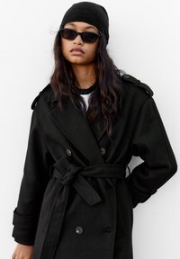 Crni dvostruko kopčani kaput s remenom, izrađen od teksturirane tkanine. Ima epaulete na ramenima i velike gumbe, uparen s crnim beaniem i sunčanim naočalama.