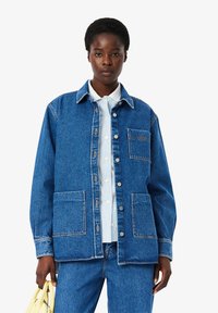 Blue denim jacket with a collar, front buttons, and two large front pockets. Worn over a light blue and white striped shirt.