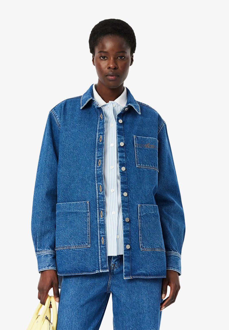 Blue denim jacket with a collar, front buttons, and two large front pockets. Worn over a light blue and white striped shirt.