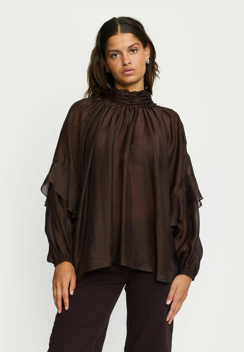 Blouse marron transparente avec un col haut à volants et des manches fluides. Présente une texture douce et une coupe ample, associée à un pantalon sombre.