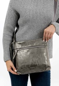 Metallic grey shoulder bag made from textured leather, features a front zip pocket and a detachable strap, held by a person.