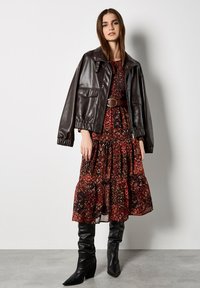 Brązowa skórzana kurtka typu cropped z elastycznymi mankietami, noszona na wzorzystą bordową sukienkę. Całość dopełniają wysokie czarne botki na obcasie.