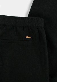 Sorte linbukser med en avslappet midje, med en baklomme og en liten oransje brodert logo. Glatt tekstur og minimalistisk design.