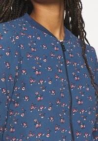 Femme portant une veste zippée bleue avec de petits motifs floraux roses et blancs, avec de longues tresses visibles sur l'épaule.