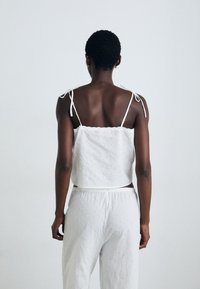 Top scurt din bumbac alb, cu bretele reglabile și detalii brodate cu flori, asortat cu pantaloni albivi, lejeri.